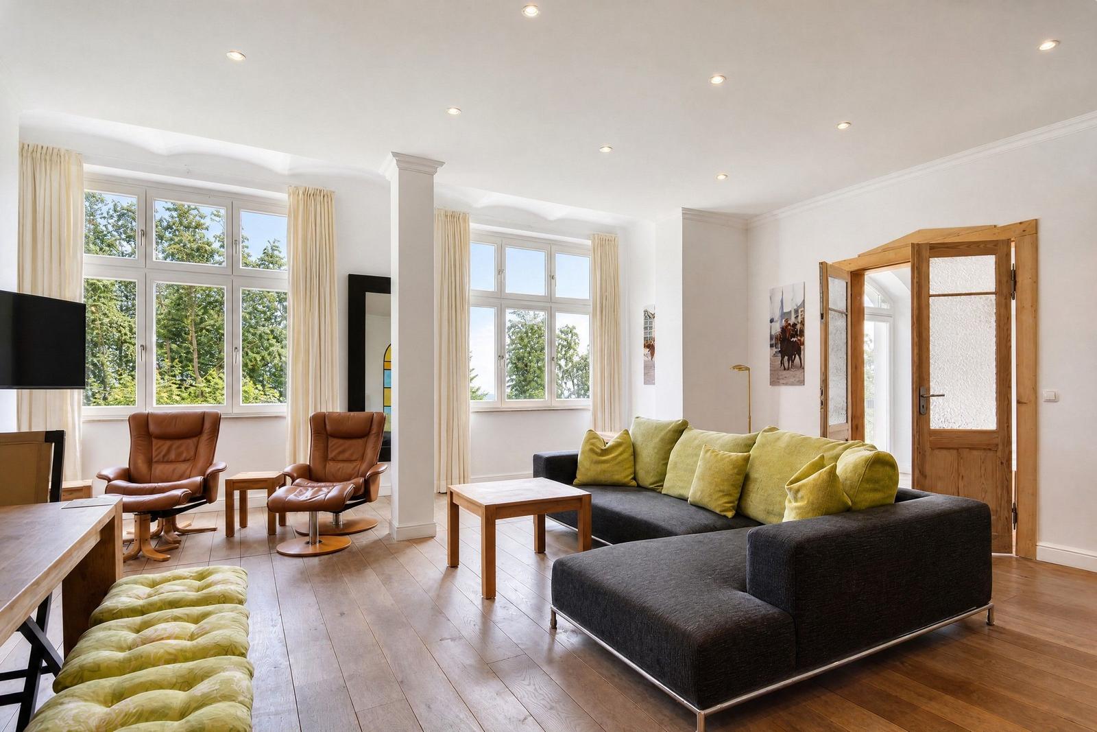 Wohnzimmer mit Sofa, Sessel und Holzboden. Große Fenster und Tür zum Garten.