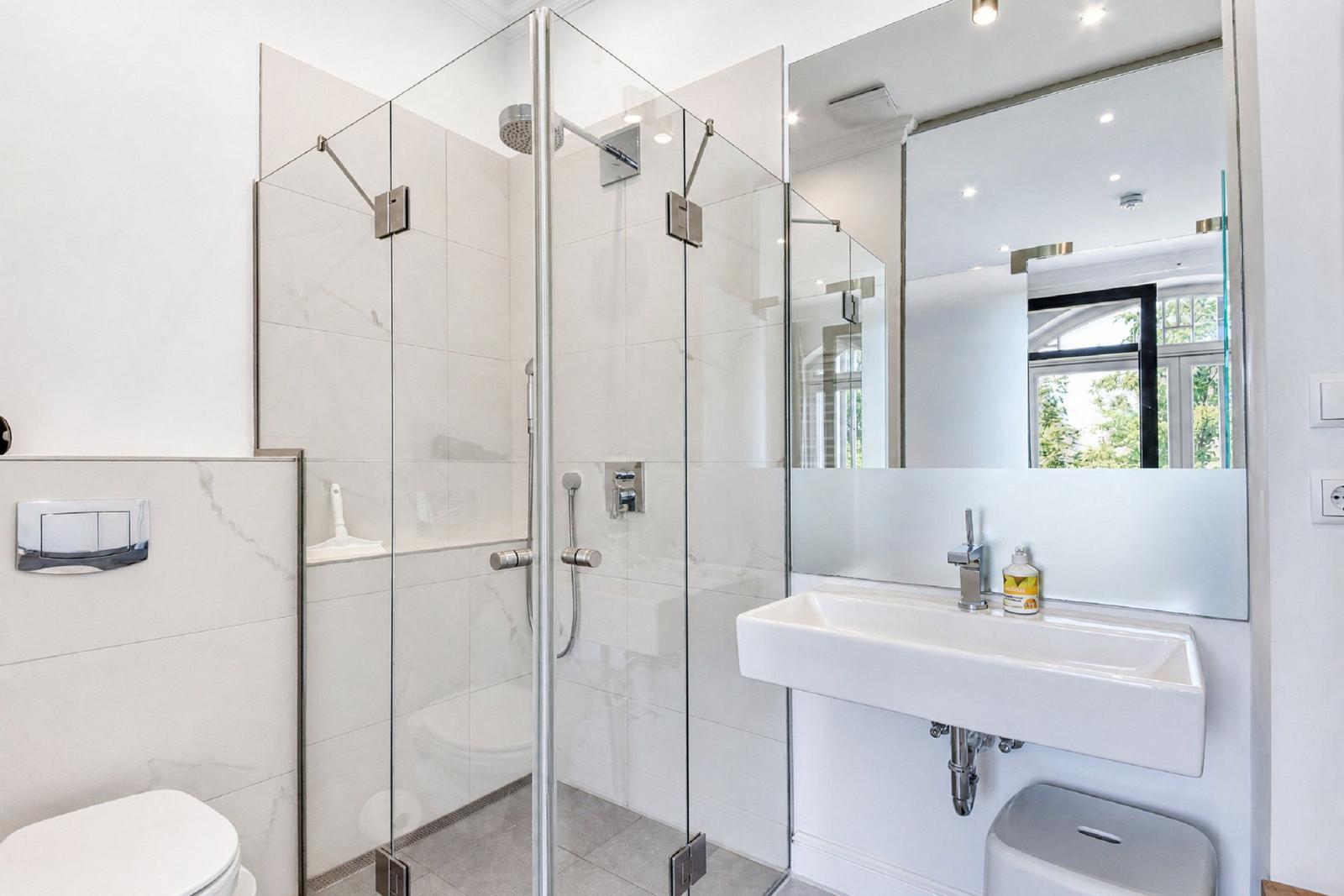 Dusche mit Glaswand, Waschbecken und Spiegel. Toilette und Fenster sichtbar.