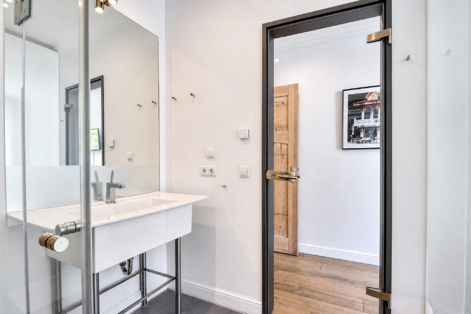 Badezimmer mit Waschbecken, Spiegel und Glasdusche. Tür führt in Wohnbereich mit Holzboden.