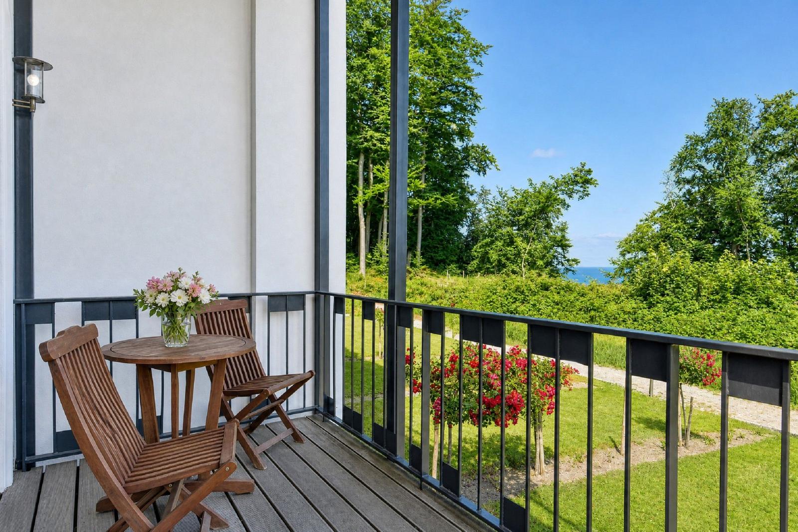 Terrasse mit Holztisch, zwei Stühlen und Blumen. Blick auf grüne Landschaft und Meer.