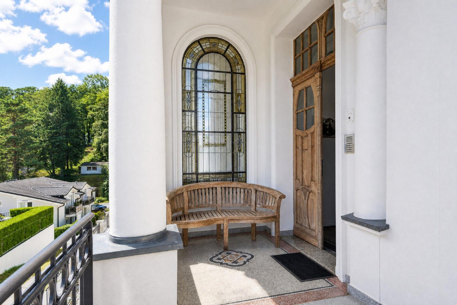 Eingangsbereich mit Holzbank, Bogenfenster und Tür. Blick auf grüne Landschaft.