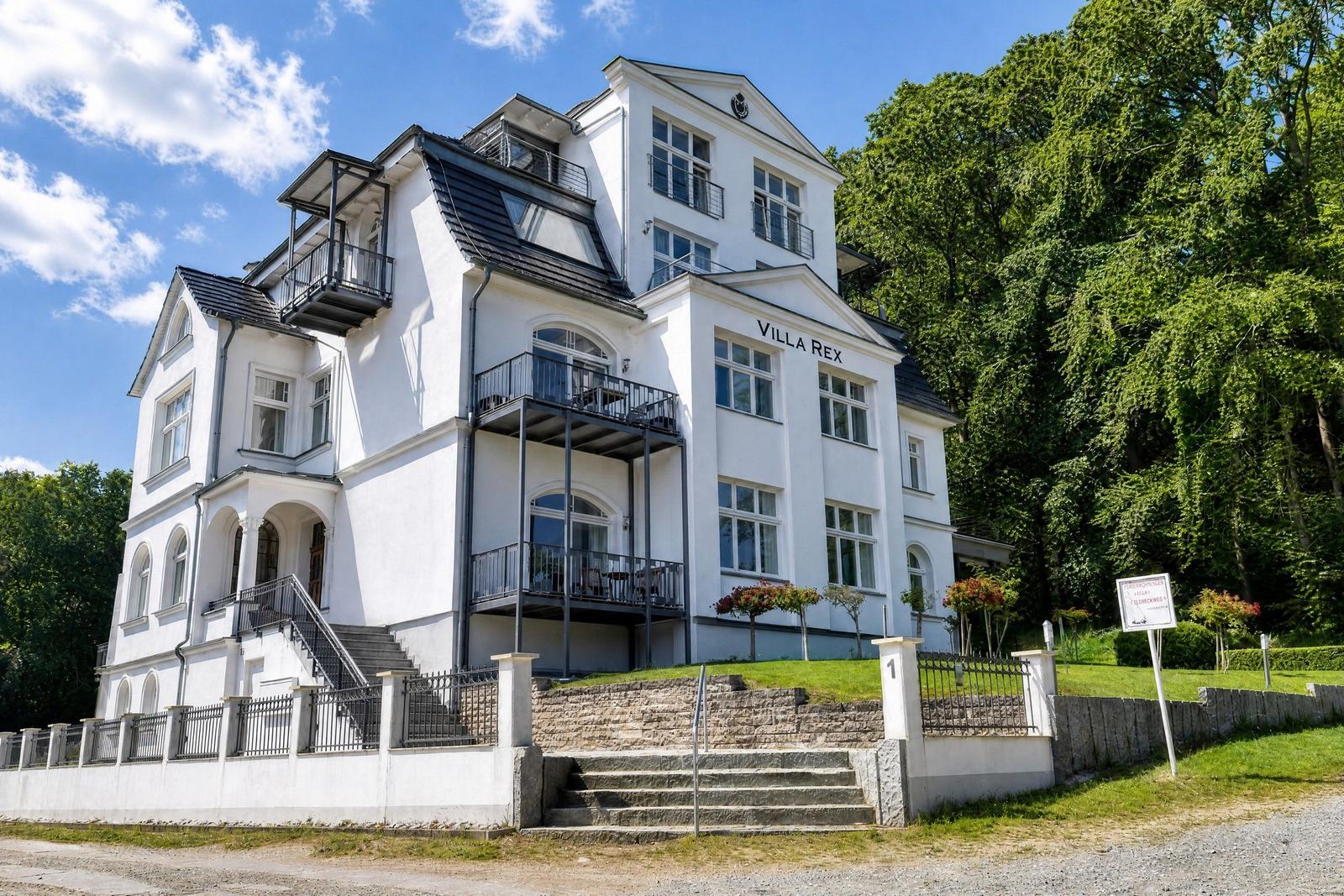 Weiße Villa mit Balkonen und Treppenhaus, umgeben von Bäumen und Grünflächen.