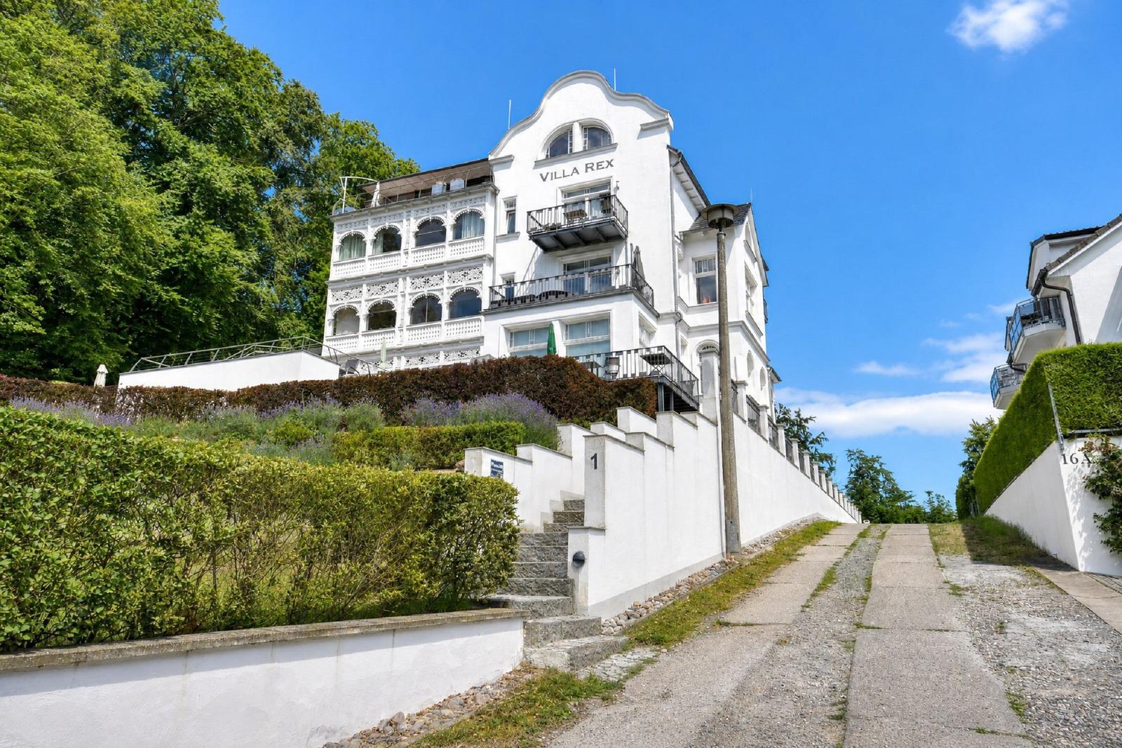 Weiße Villa mit Balkonen und Gartenanlage an steilem Hang.