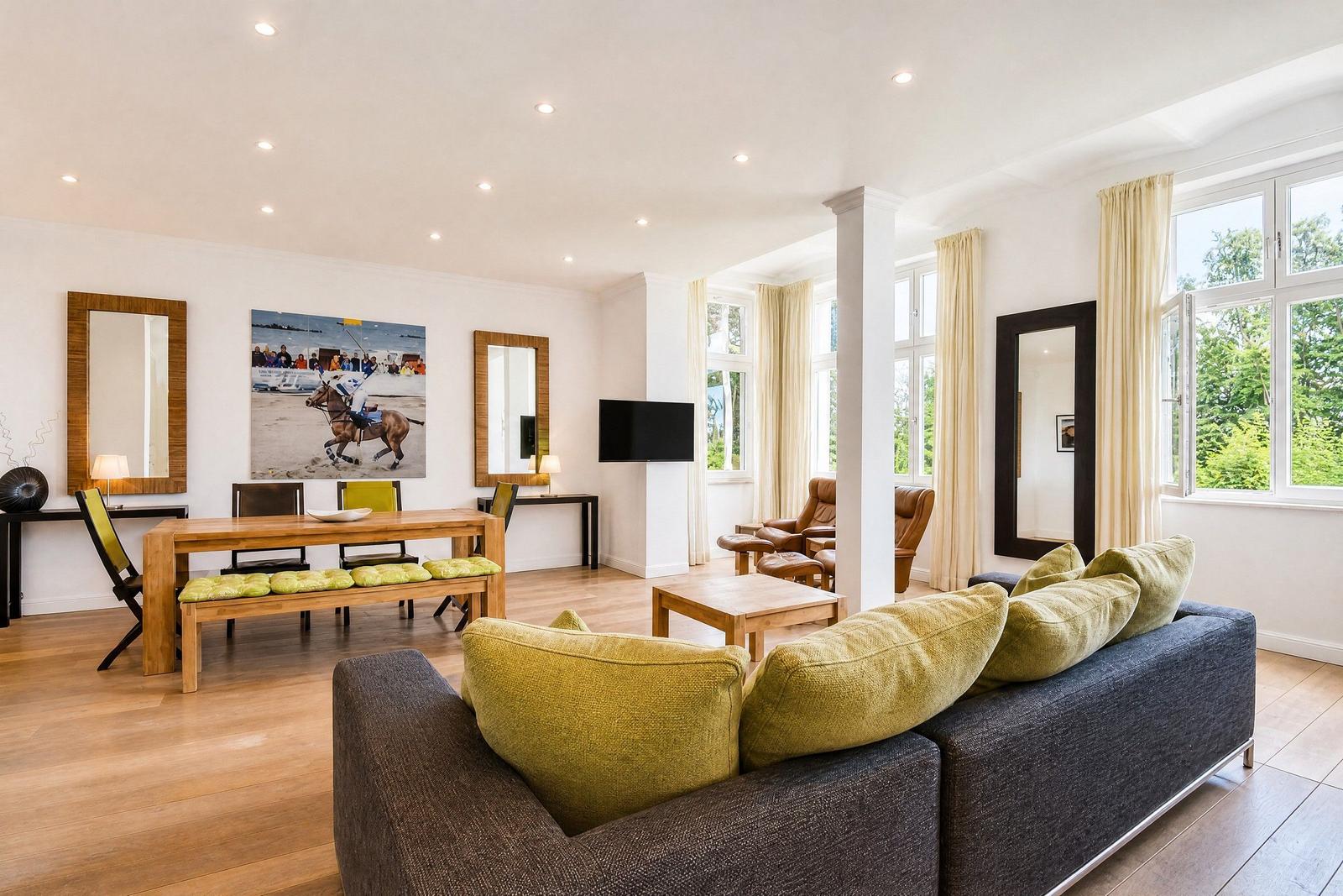 Wohnzimmer mit Sofa, Esstisch und großen Fenstern. Wandbild und Spiegel sichtbar.