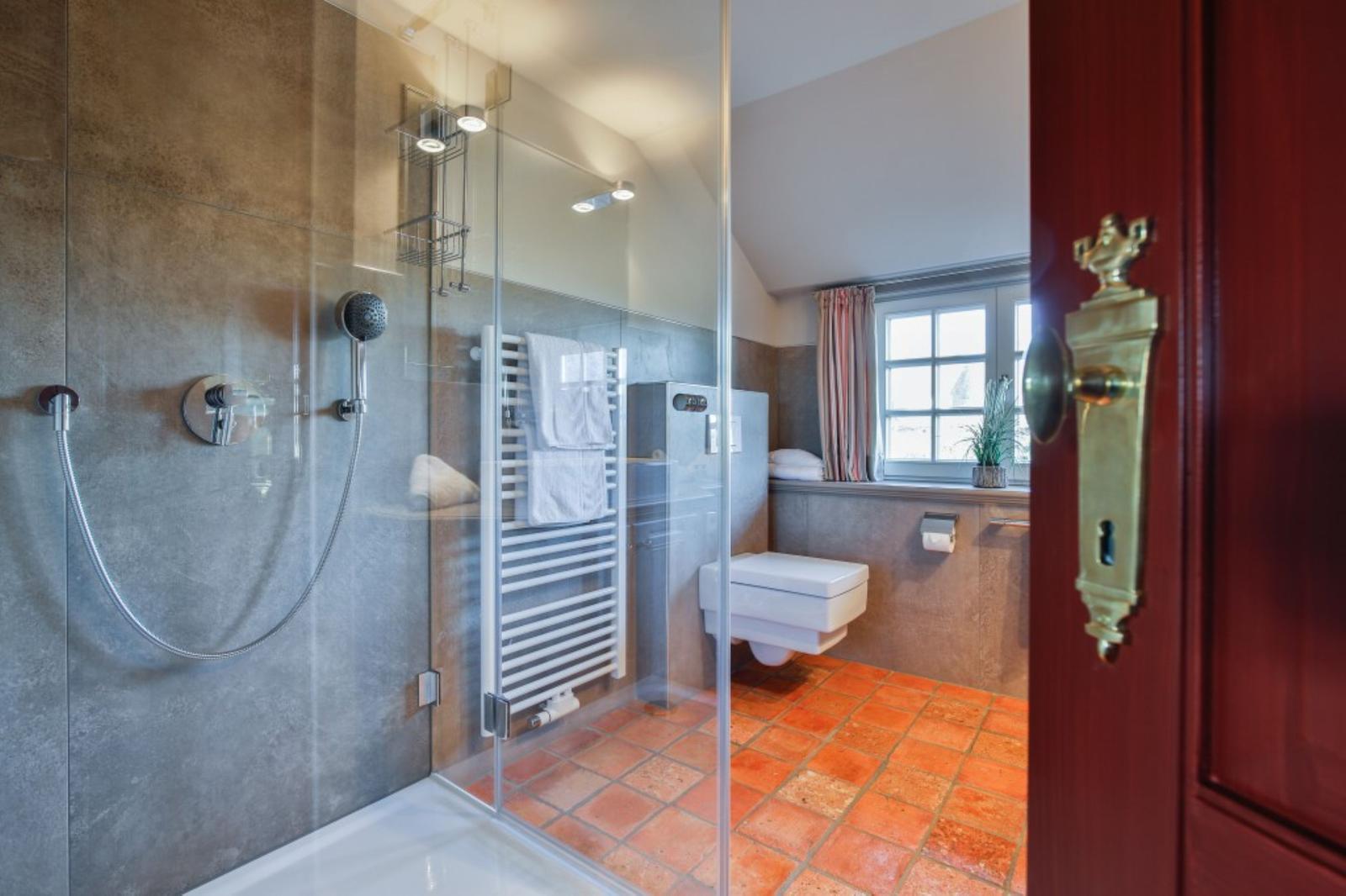 Badezimmer mit Dusche, Toilette und Fenster. Terrakottafußboden und Heizkörper.