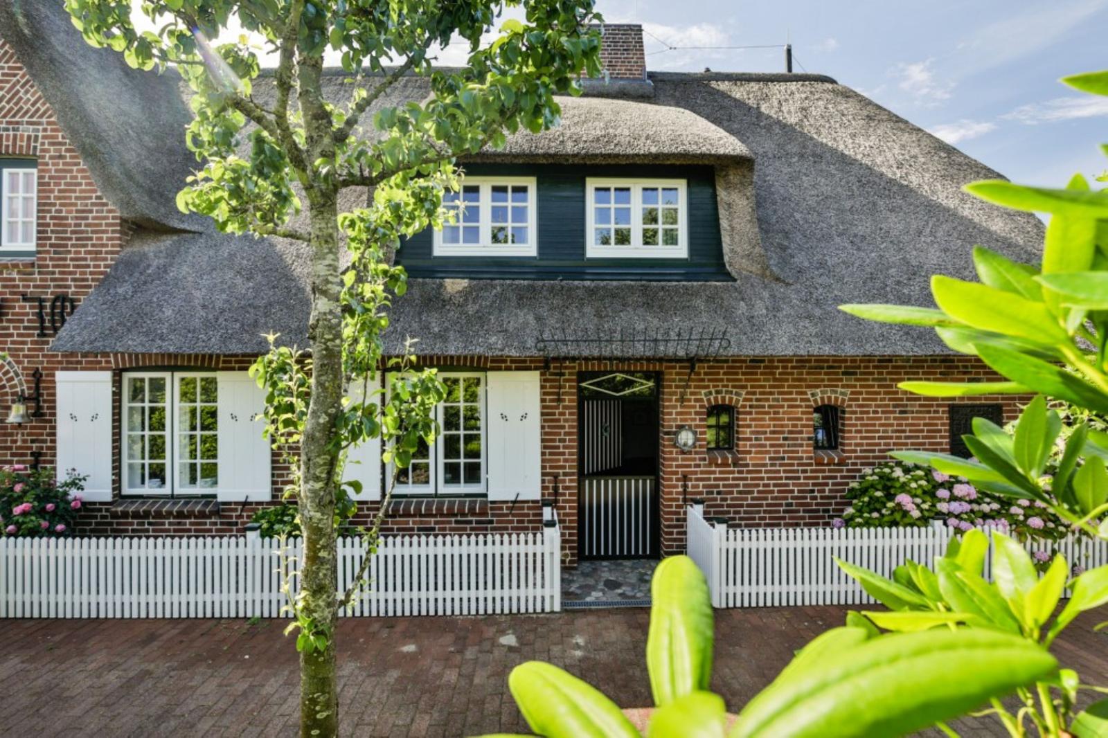 Haus mit Strohdach, rotem Backstein, weißer Tür und Fensterläden, umgeben von Grün und weißen Zäunen.