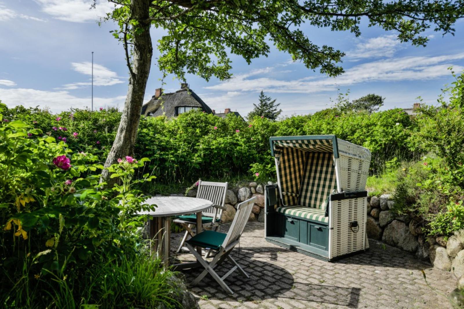 Terrasse mit Strandkorb, Tisch und Stühlen umgeben von grünen Pflanzen und einem Baum.