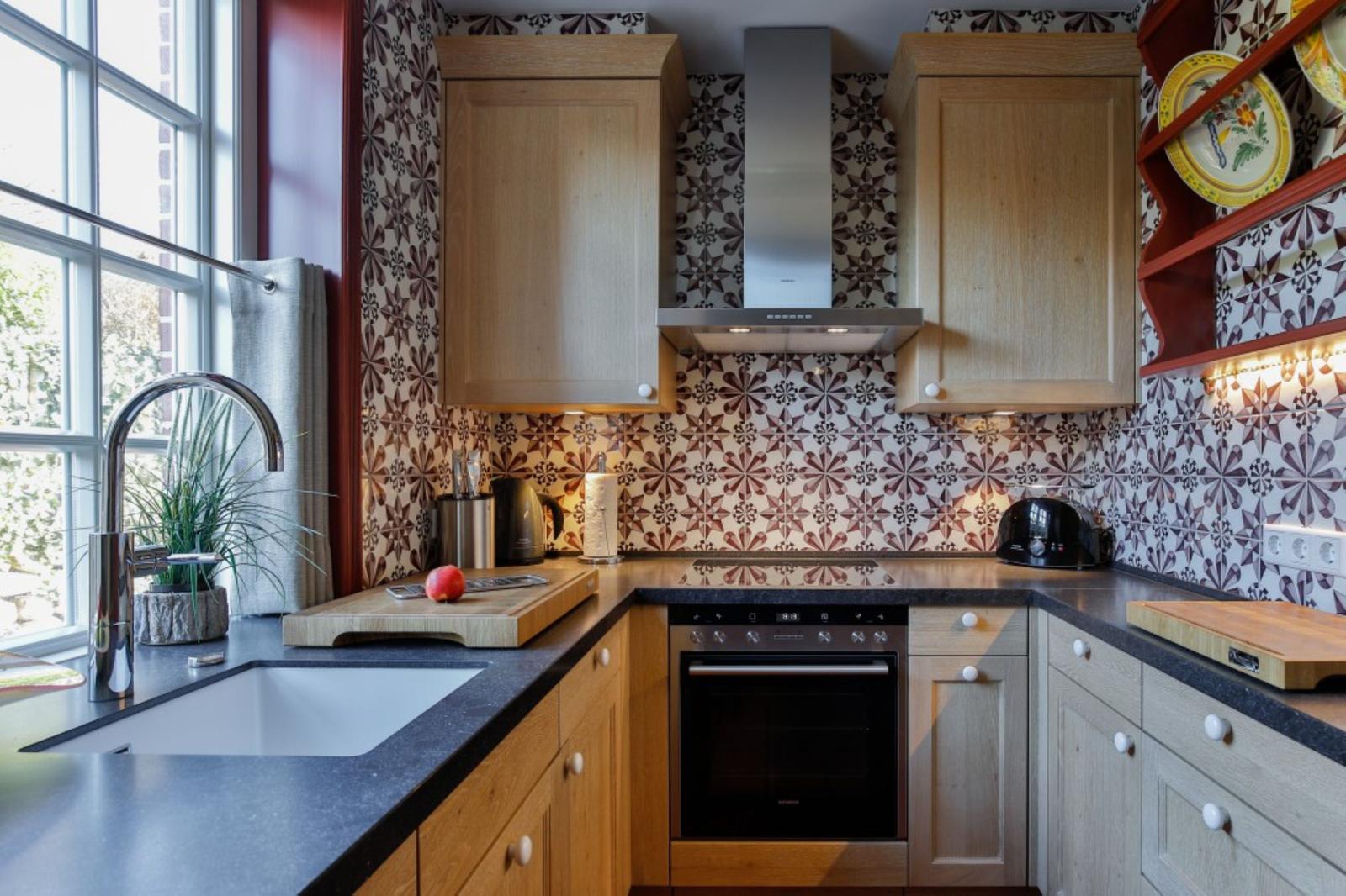 Küche mit Holzschränken, Mosaikwand und Edelstahlspüle. Fenster, Arbeitsfläche und Ofen sichtbar.
