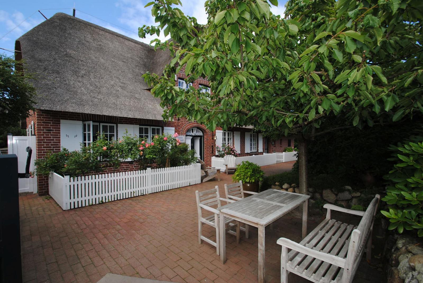 Haus mit strohgedecktem Dach, weißer Zaun und Gartenmöbel auf gepflastertem Platz.