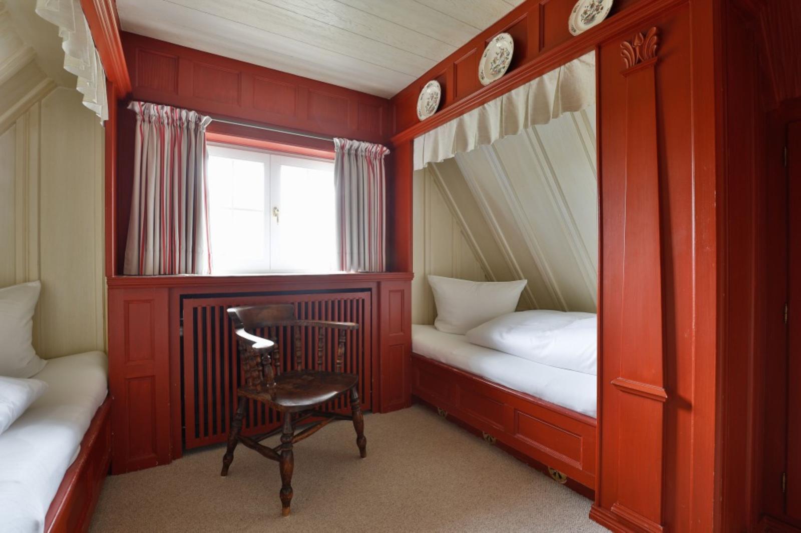 Zimmer mit zwei Betten, Holzstuhl und Fenster mit Vorhängen.