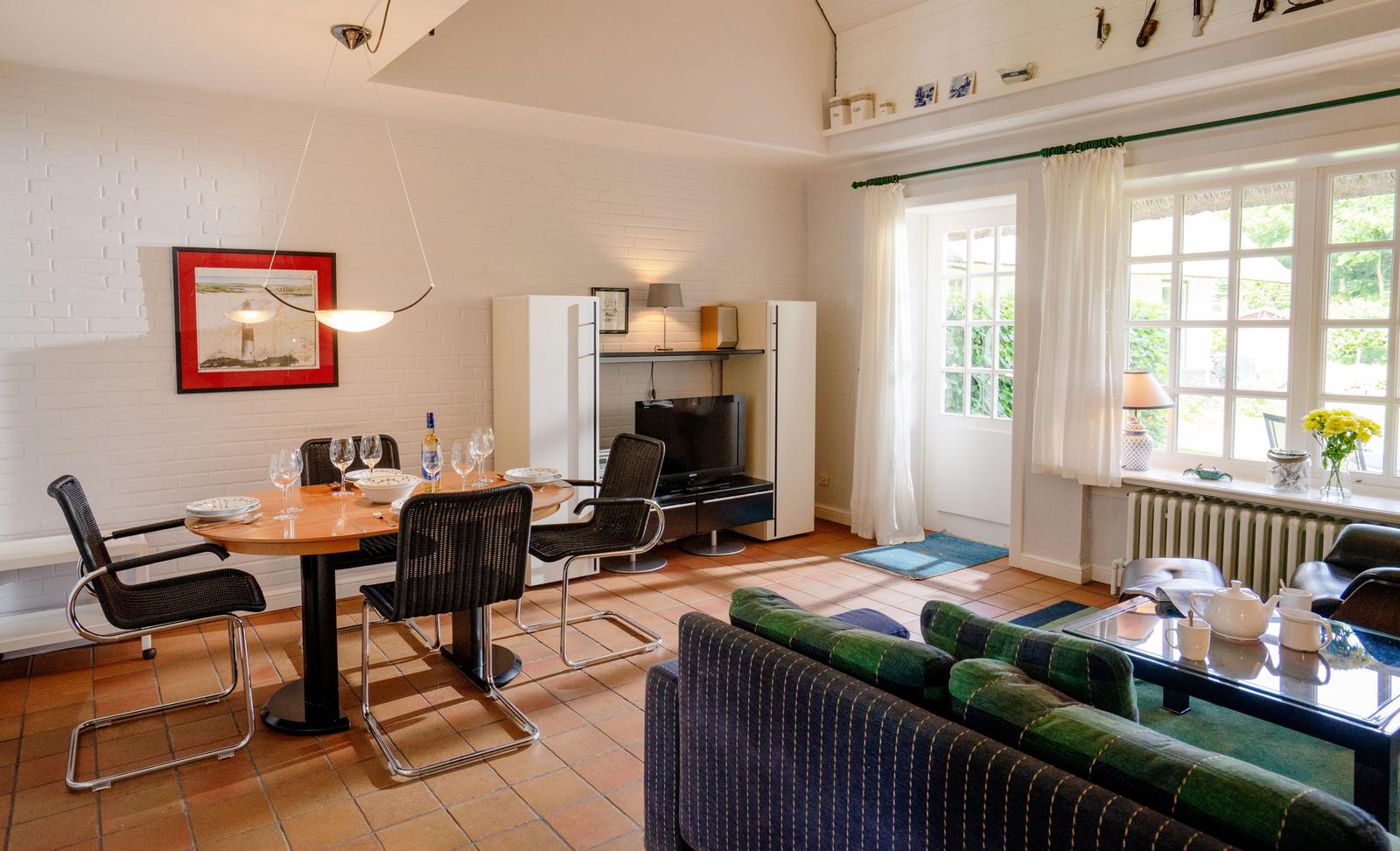 Wohn- und Essbereich mit Tisch, Stühlen, Sofa und Fernseher. Fenster mit Vorhängen und Blick nach draußen.
