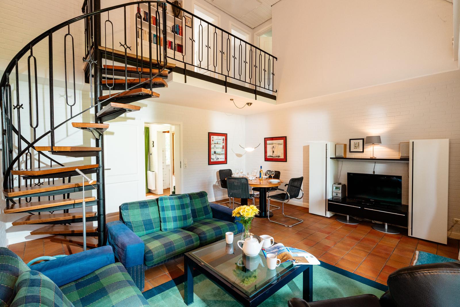 Wohnzimmer mit Treppe, Sofa, Couchtisch und Fernseher. Terrakottaboden und weiße Wände.