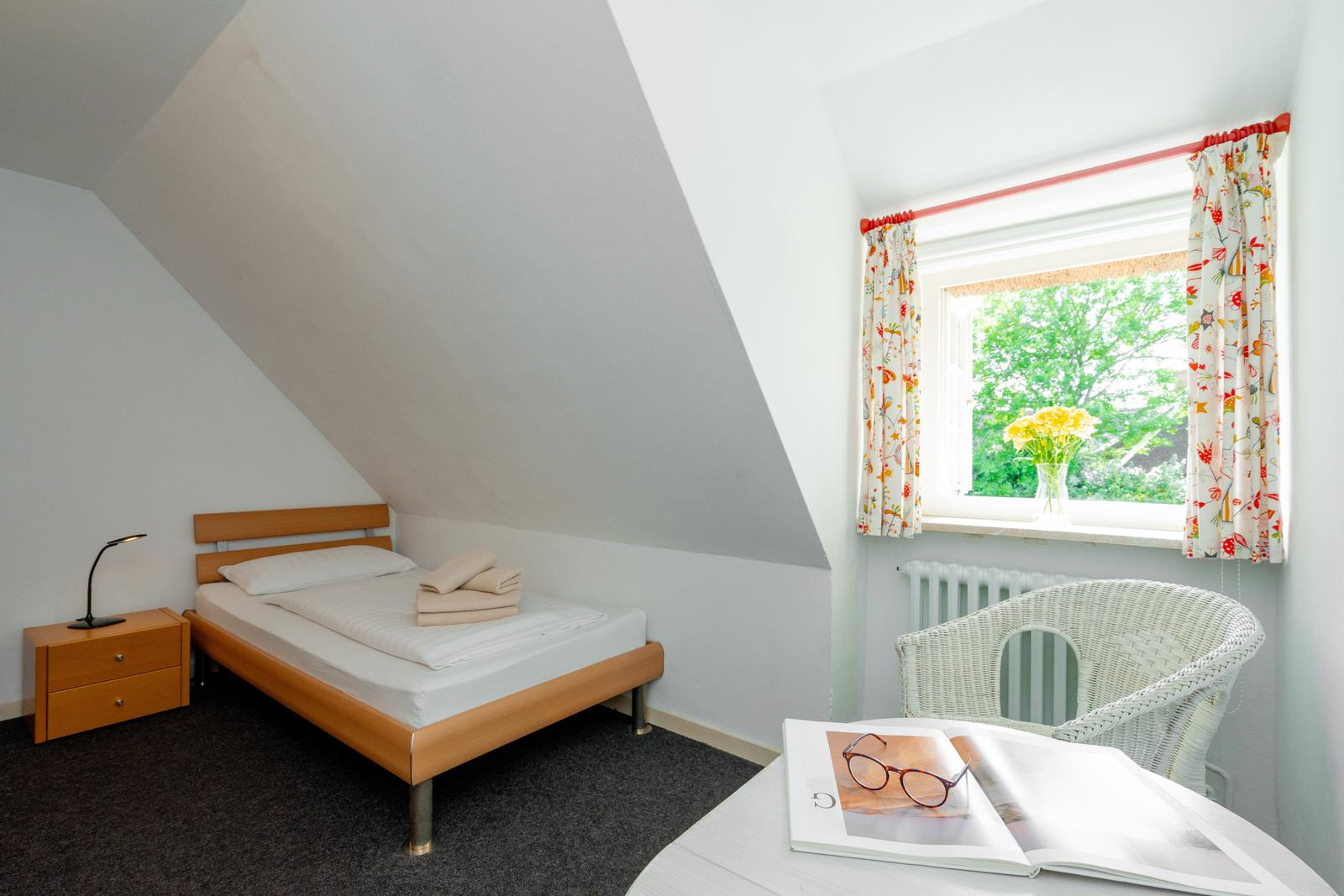 Ein Schlafzimmer mit Dachschrägen, Bett, Nachttisch und Fenster mit Blumen.