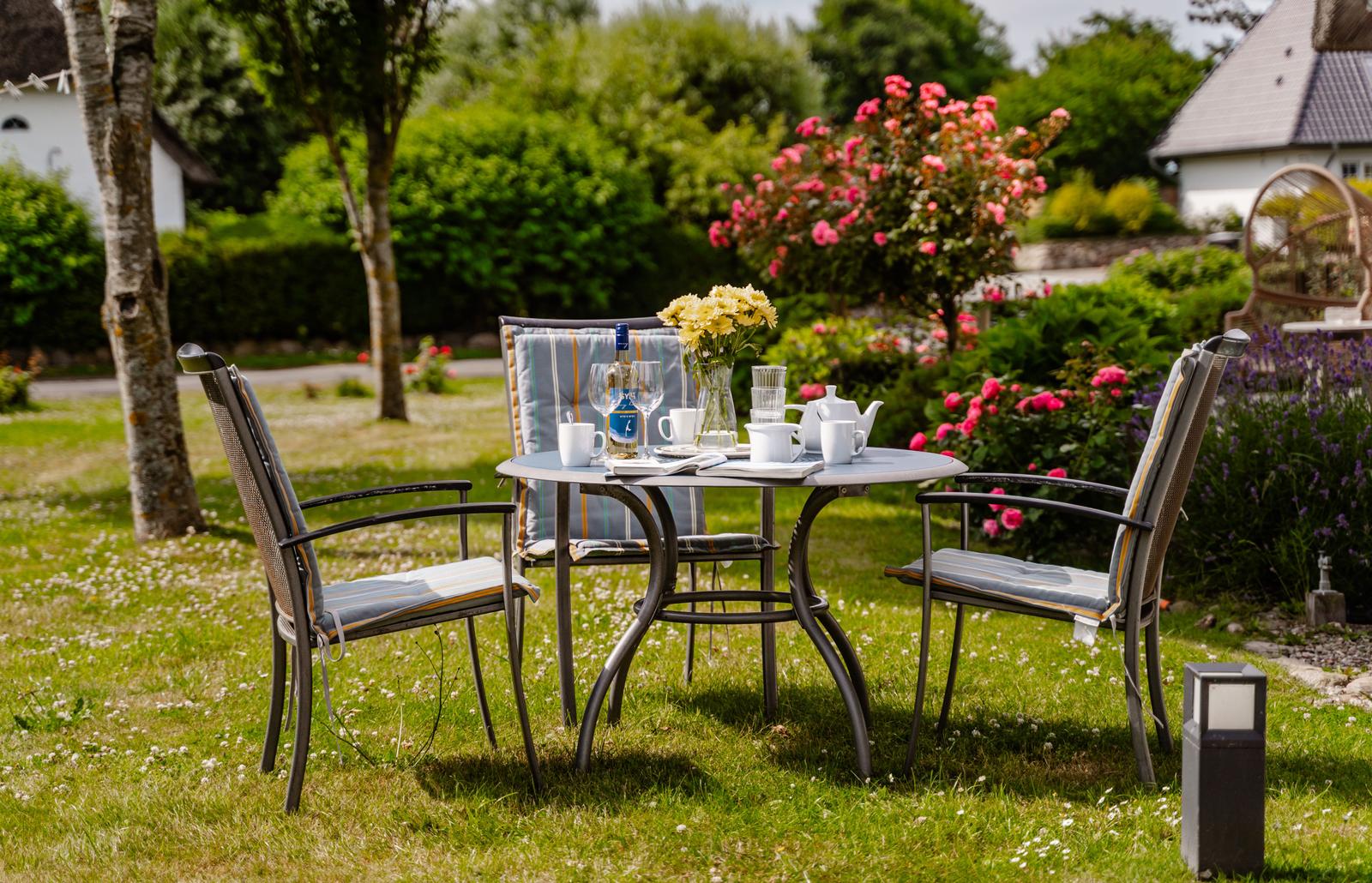 Garten-Tisch mit Stühlen, Blumen und Getränken. Hintergrund: Blumenbeete und Haus.