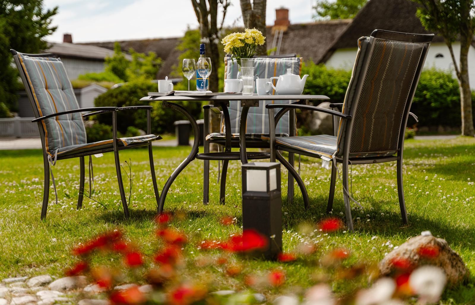 Gartenbereich mit Tisch, Stühlen und Blumen. Hintergrund: Haus mit Dach.