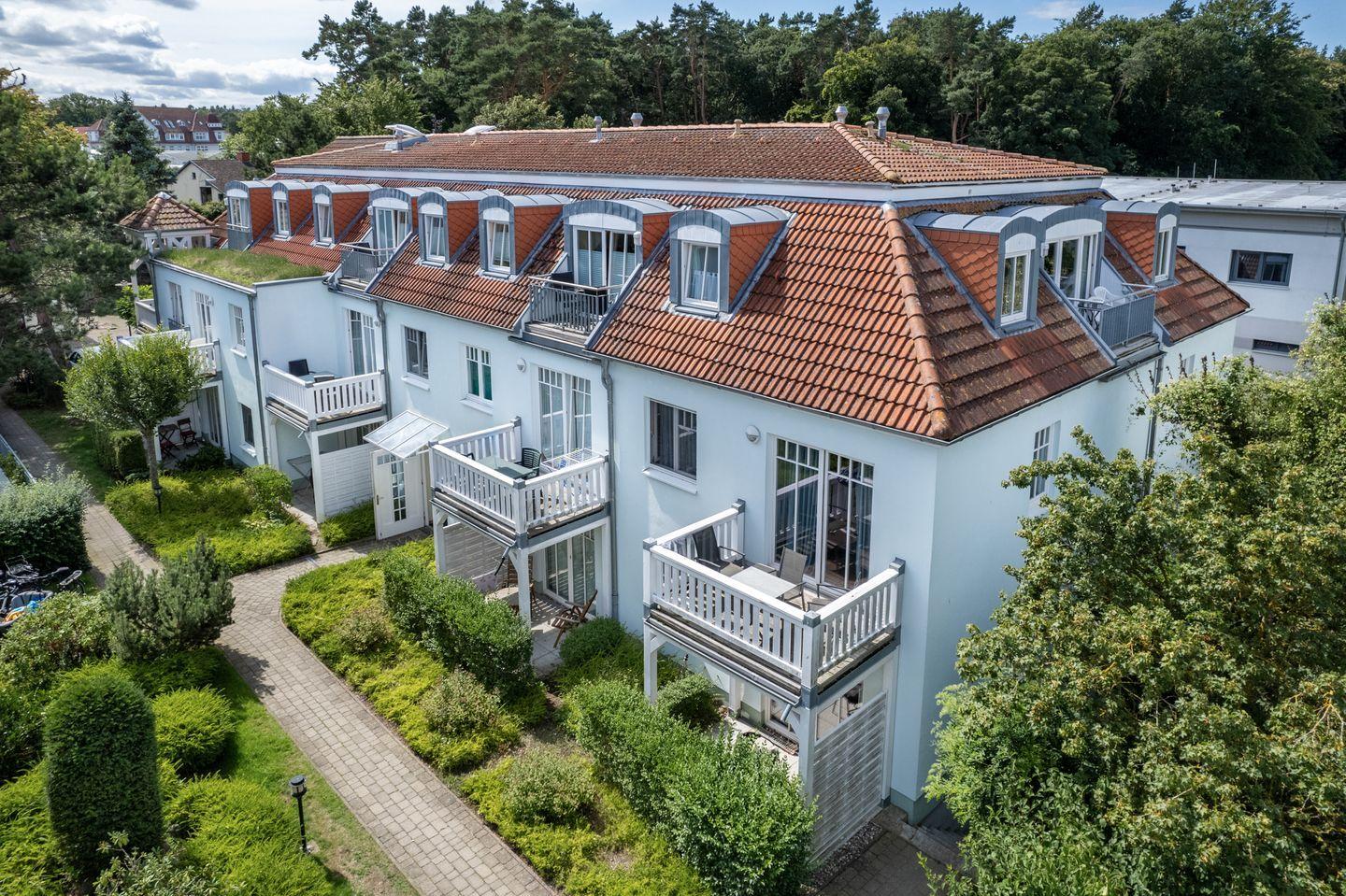 Ferienhaus mit mehreren Balkonen und rotem Ziegeldach. Umgeben von Grünflächen und Bäumen.