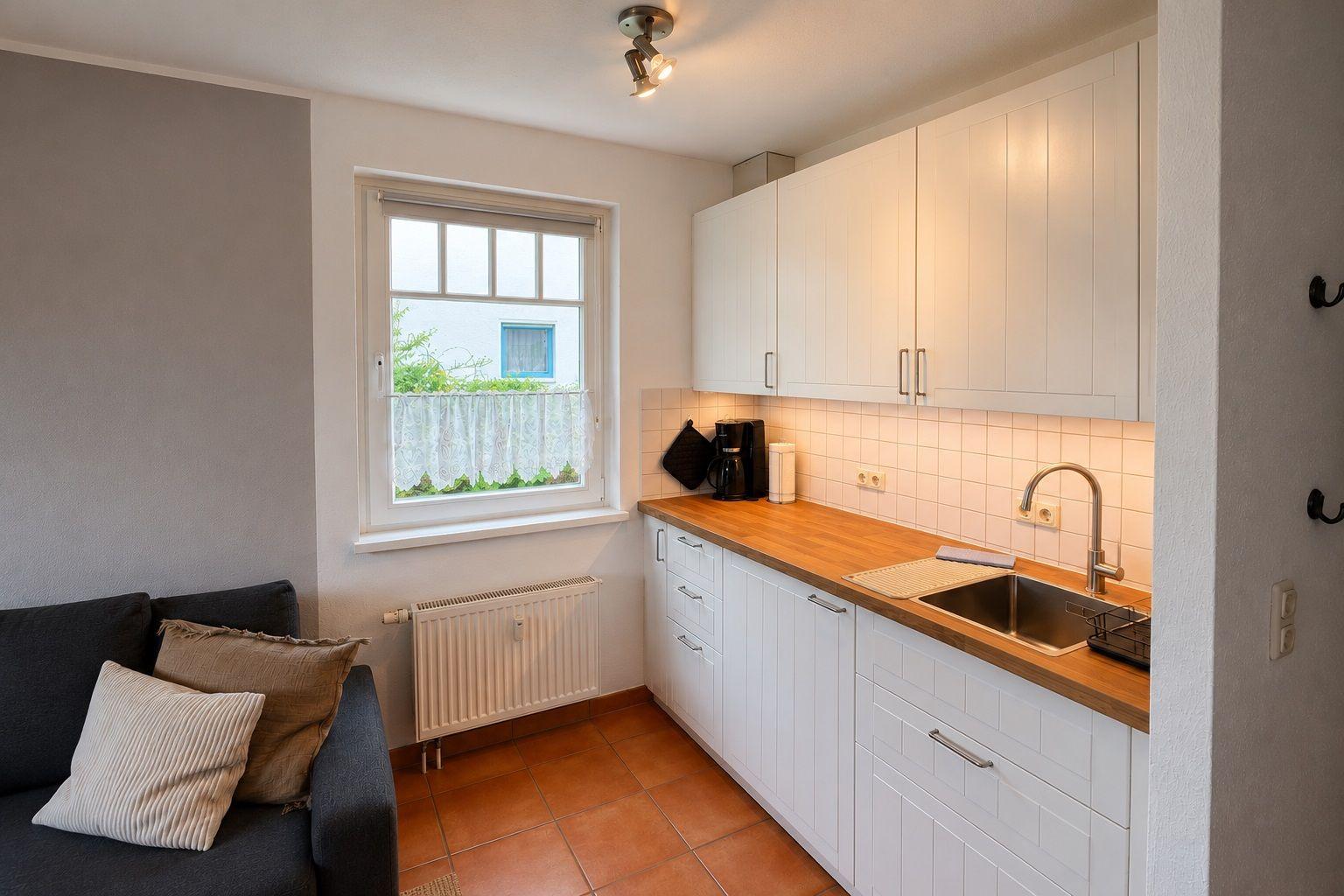 Küche mit weißen Schränken, Holz-Arbeitsplatte und Fenster. Links Wohnbereich mit Sofa.