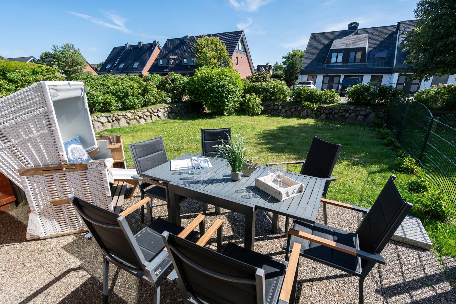 Terrasse mit Tisch, Stühlen und Liegestuhl. Hintergrund: Grasfläche und Häuser.