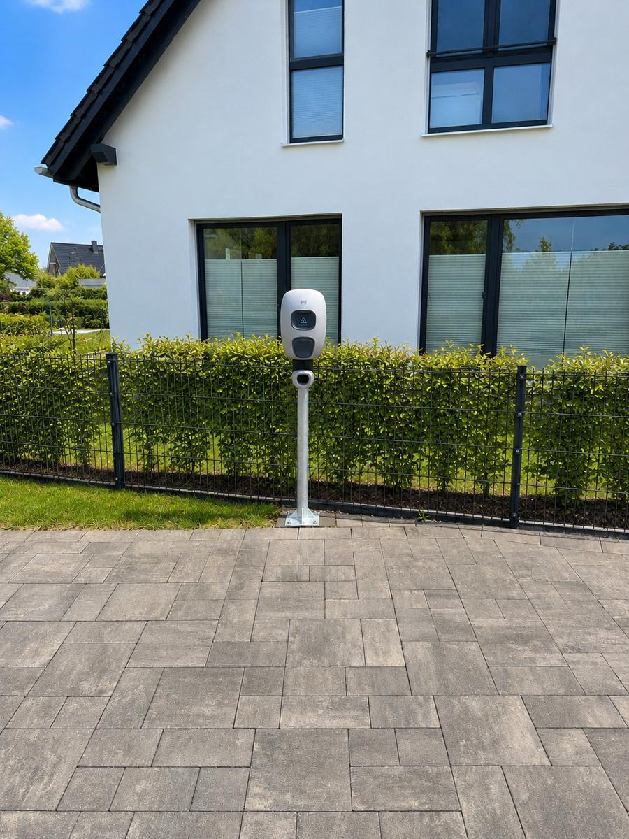 Ladestation auf gepflastertem Platz vor modernem Haus mit Hecke und Fenstern.
