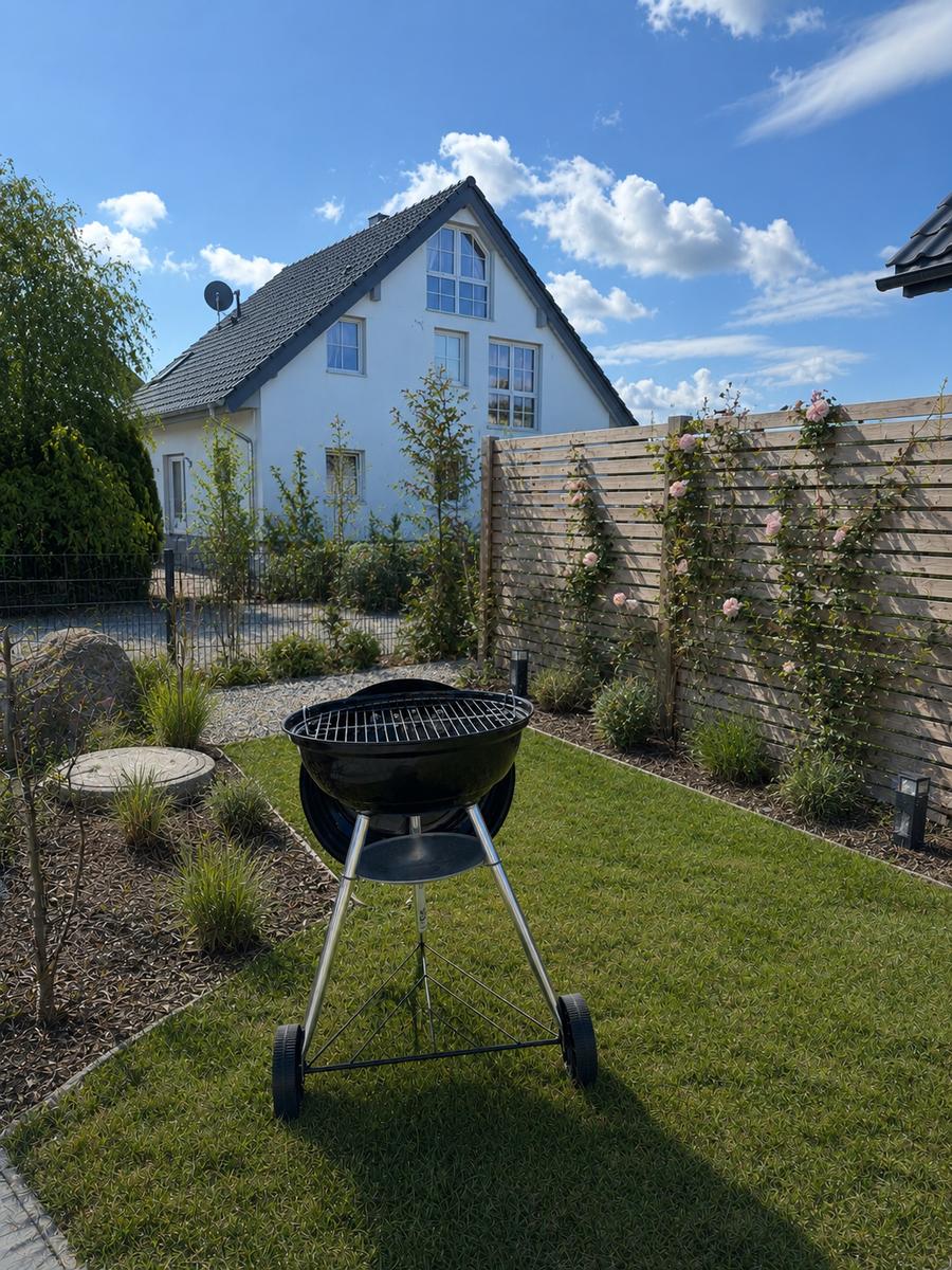 Garten mit Grill, Rasen und Holzzaun. Im Hintergrund das weiße Haus.