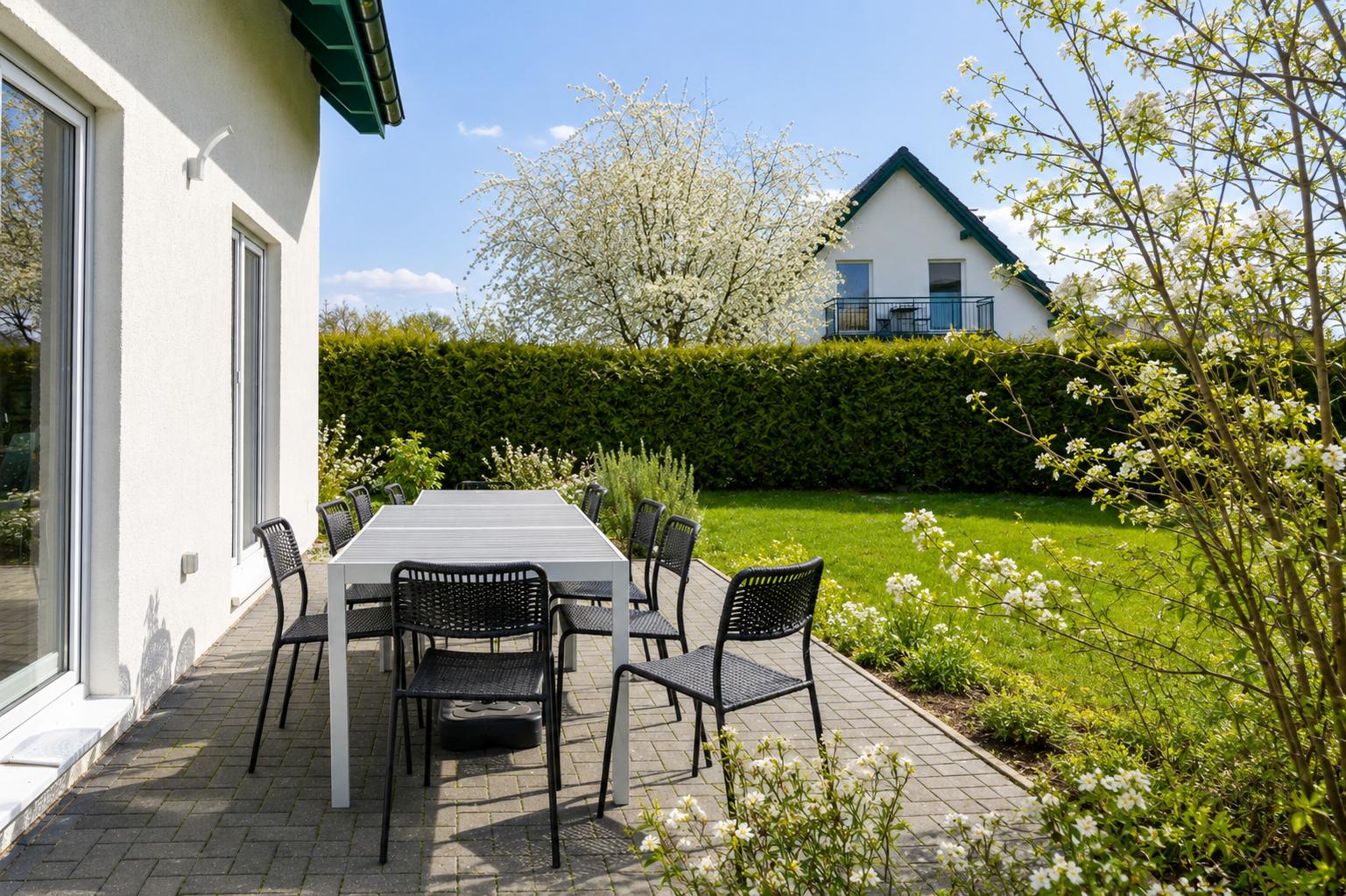 Terrasse mit Tisch und Stühlen, grüner Rasen, blühende Sträucher und weißes Haus im Hintergrund.