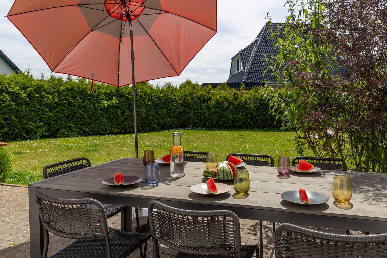 Garten-Tisch mit Sonnenschirm und Wassermelonen. Hintergrund: Grasfläche und Hecke.