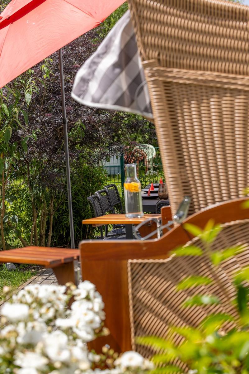 Terrasse mit Rattanmöbeln, rotem Sonnenschirm und Blumenbeet im Hintergrund.