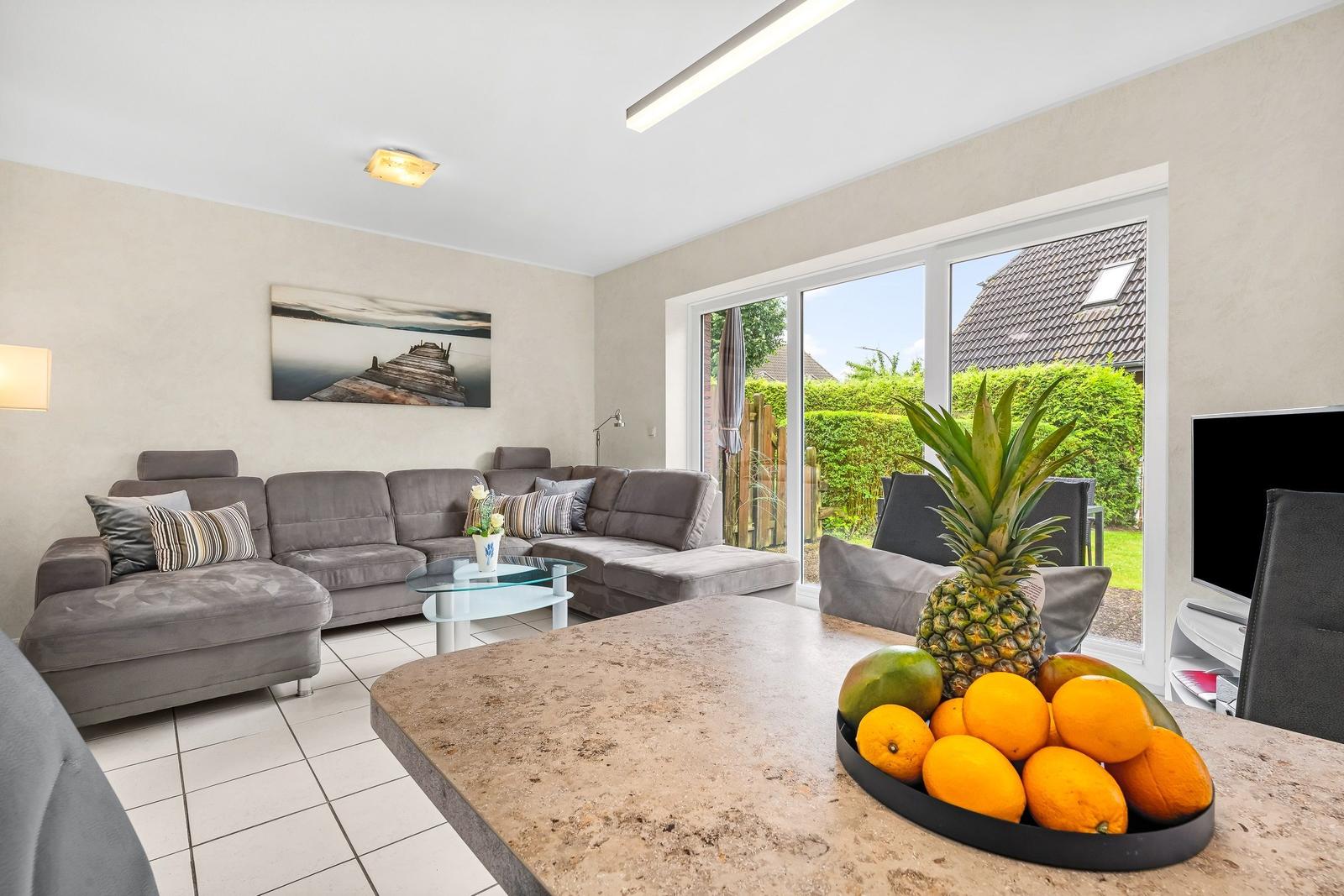 Wohnzimmer mit grauem Sofa, Glas-Tisch und Blick auf Garten. Obstschale auf Küchenarbeitsplatte.