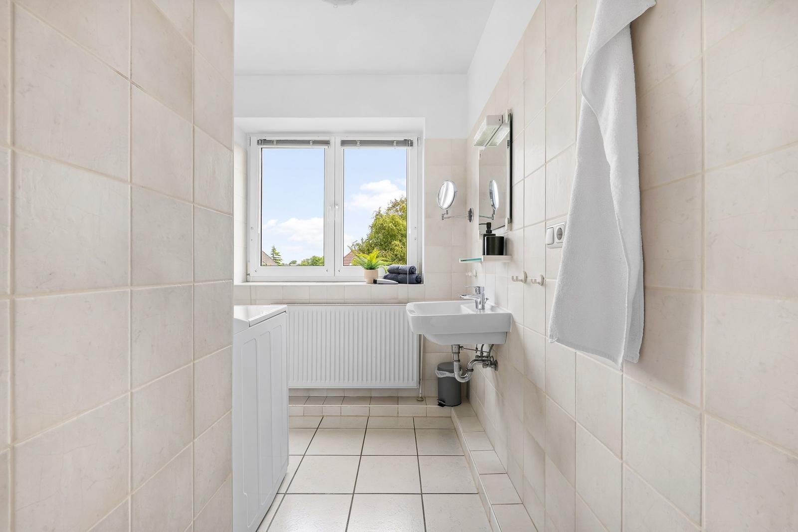 Badezimmer mit Waschmaschine, Waschbecken und Fensterblick auf Grün.