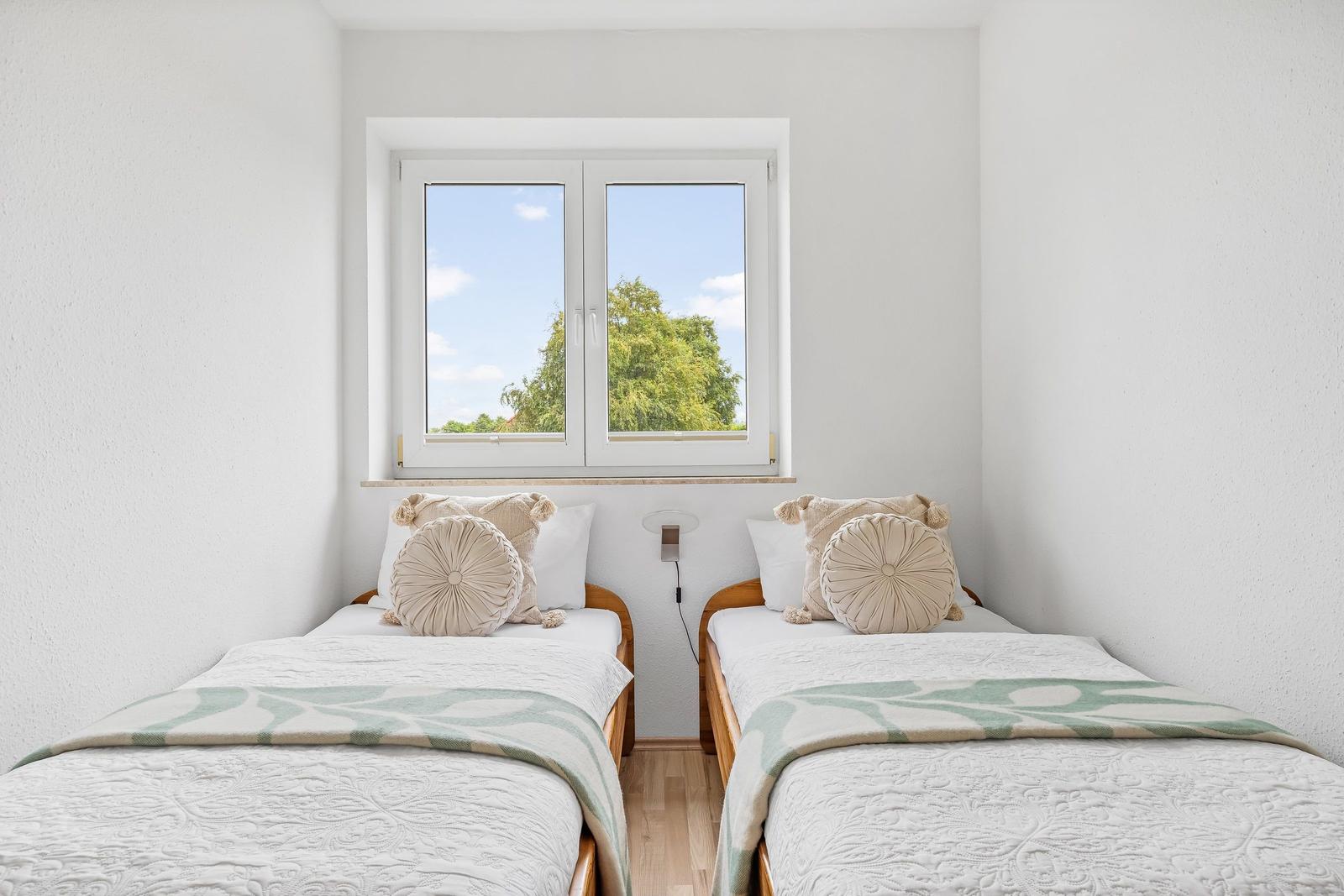 Zwei Einzelbetten mit Kissen und Decke unter einem Fenster mit Blick auf Bäume.