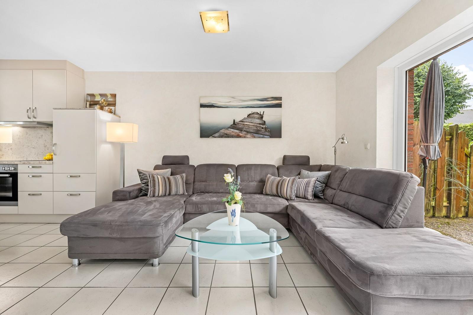 Großer Wohnbereich mit grauem Ecksofa, Glas-Tisch und Küchenbereich im Hintergrund.