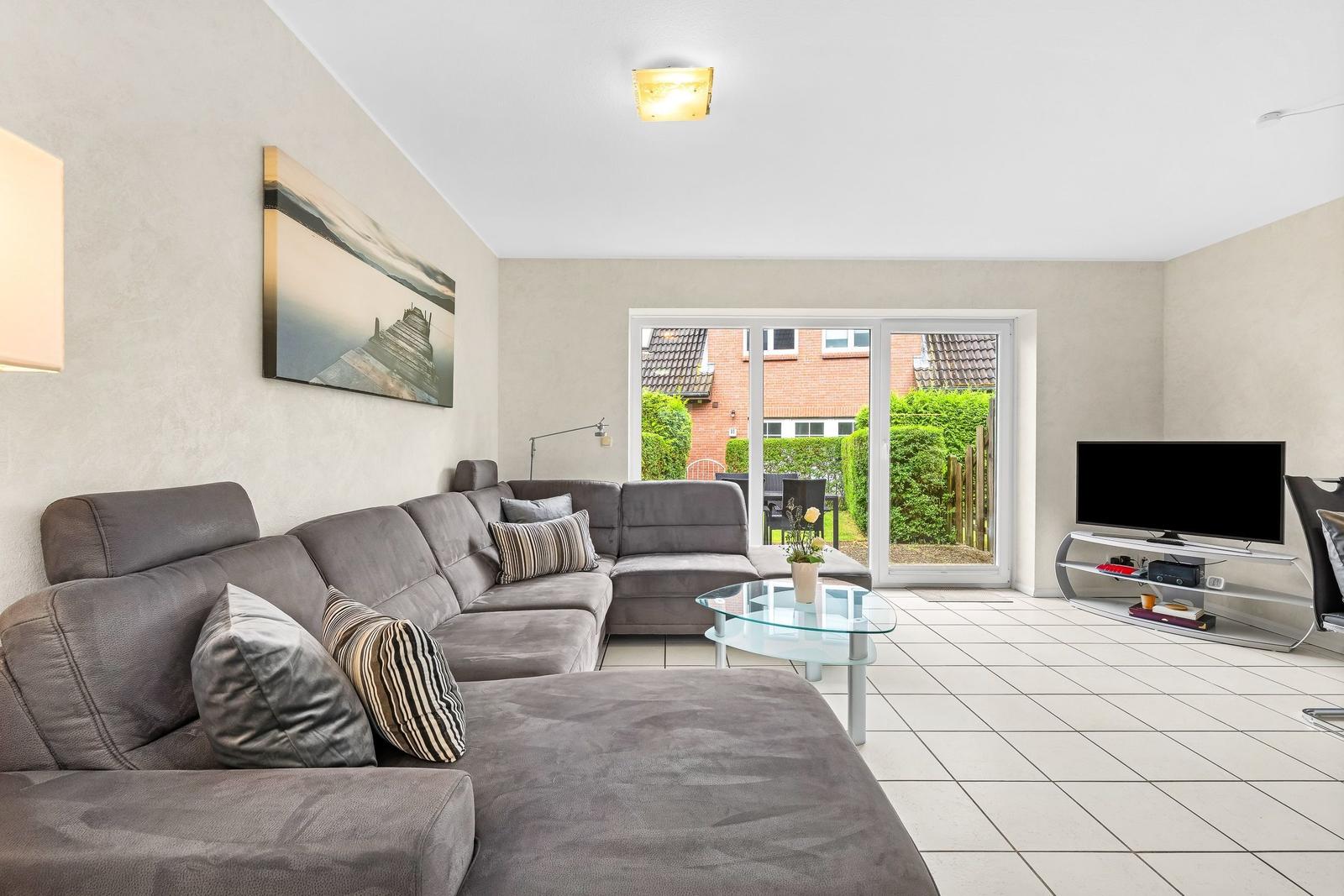 Ein Wohnzimmer mit grauem Sofa, Glas-Tisch und TV. Durch Glastür ist Garten sichtbar.