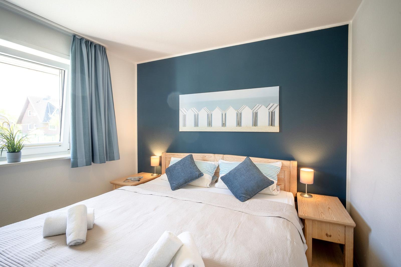 Schlafzimmer mit Bett, Nachttischen und Wandbild. Fenster mit Vorhang und Pflanze.