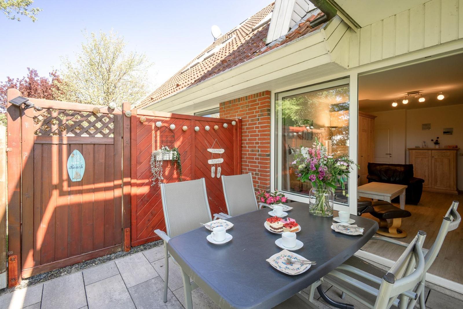 Terrasse mit Tisch, Stühlen und Blumen. Durchgehendes Fenster zeigt Wohnraum.