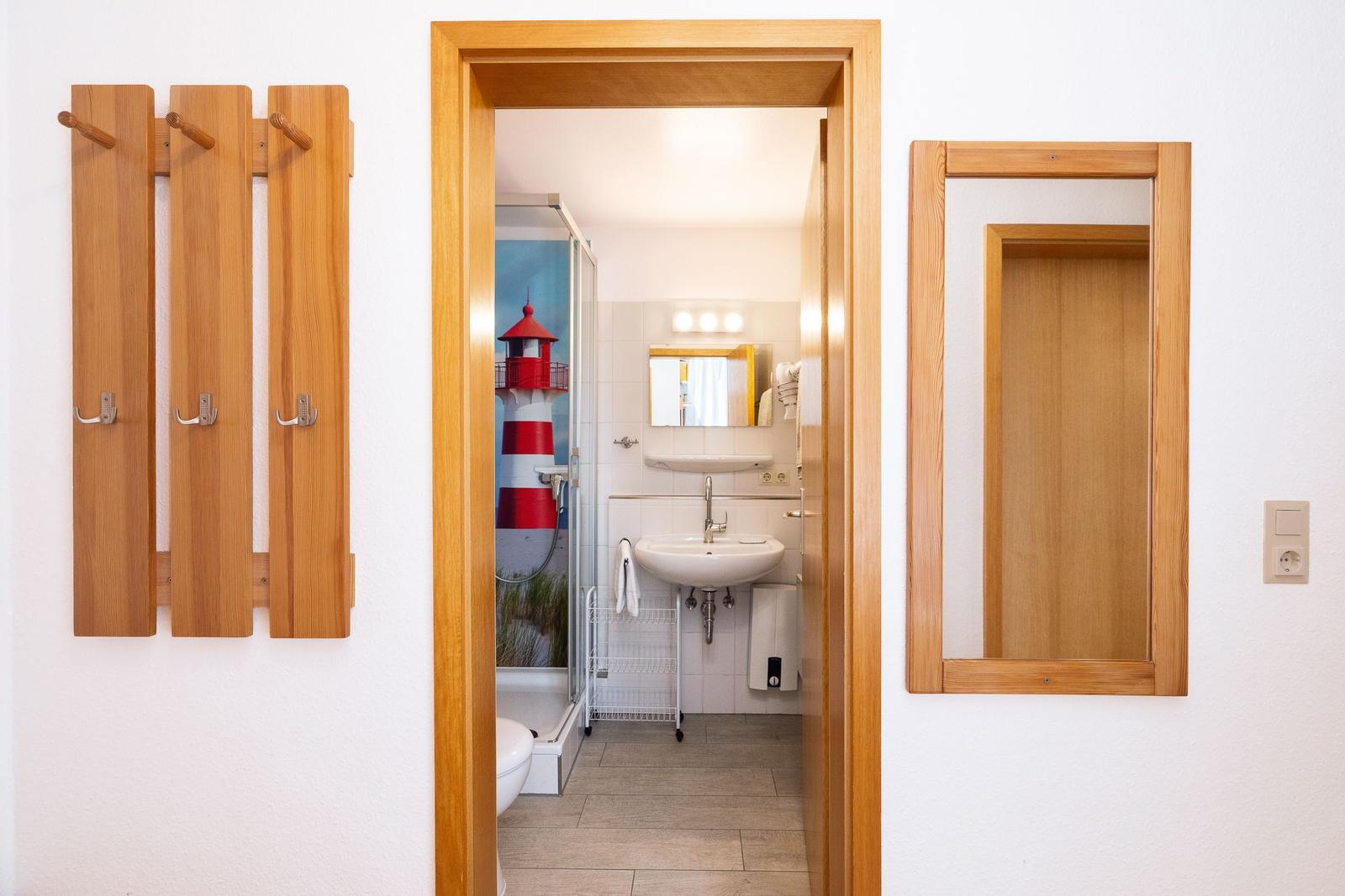 Badezimmer mit Dusche, Waschbecken und Spiegel. Holzrahmen und Garderobe sichtbar.