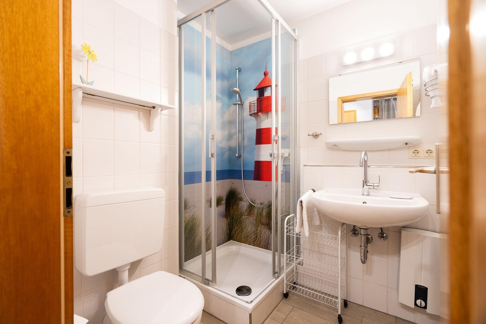 Badezimmer mit Dusche, Waschbecken und Toilette. Duschwand zeigt Leuchtturm-Motiv.