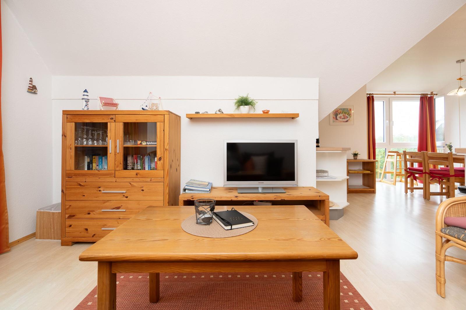 Wohnzimmer mit Holzmöbeln, Fernseher und Blick in die Küche.