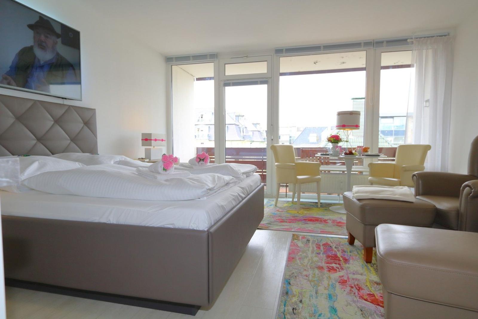 Schlafzimmer mit Bett, Fernseher und Balkonansicht. Sitzbereich mit Stühlen und Teppich.