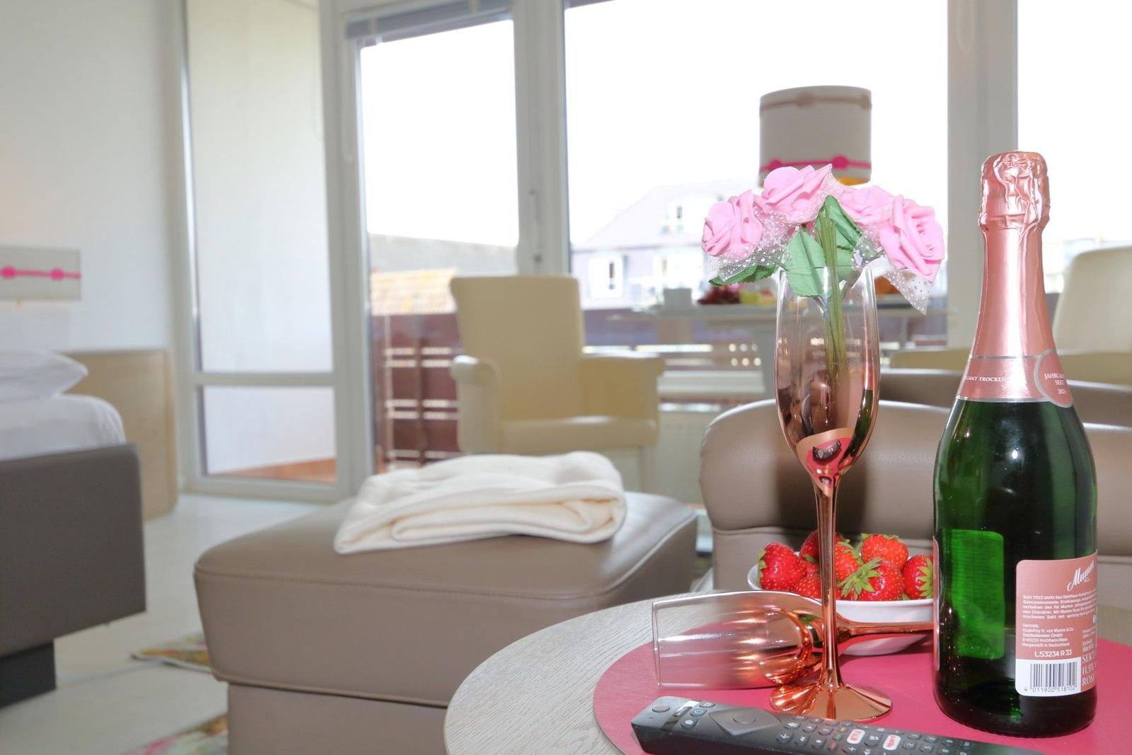 Auf dem Tisch steht eine Flasche Sekt, ein Glas und Erdbeeren. Im Hintergrund ist ein Balkon mit Sitzmöbeln.