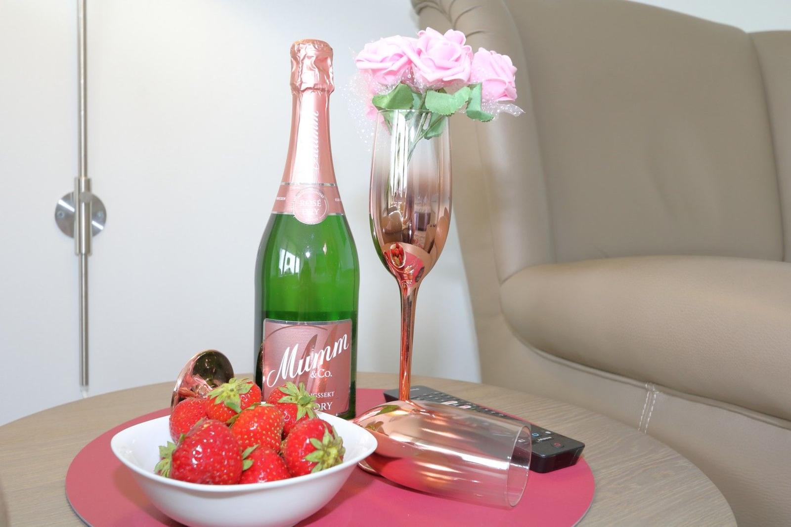 Auf einem Tisch stehen eine Flasche Sekt, ein Glas, eine Schale Erdbeeren und rosa Rosen.