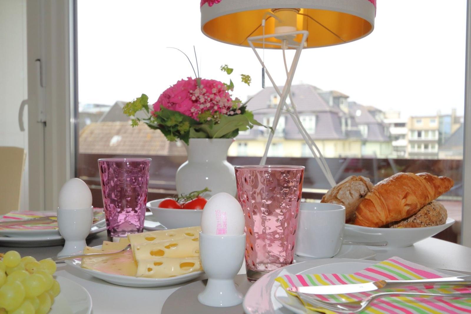 Frühstückstisch mit Käse, Eiern, Croissants und Blumen vor Fenster.