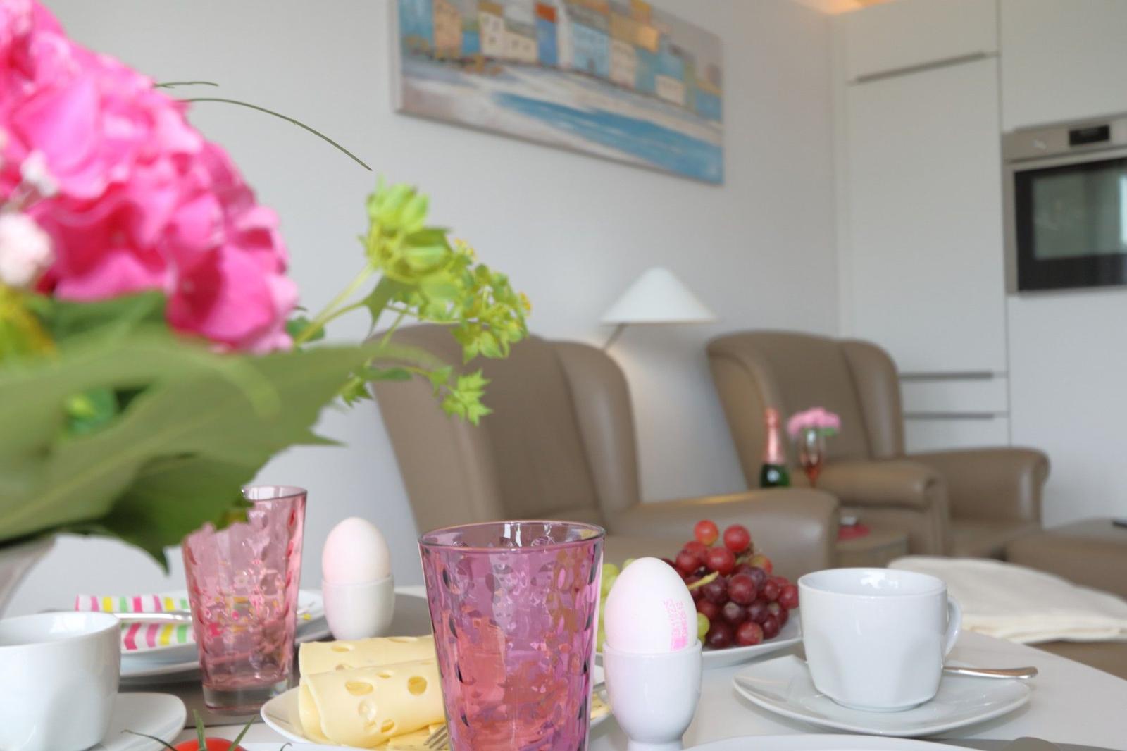Frühstückstisch mit Blumen, Obst und Getränken. Im Hintergrund Sitzgelegenheiten und Küchenbereich.
