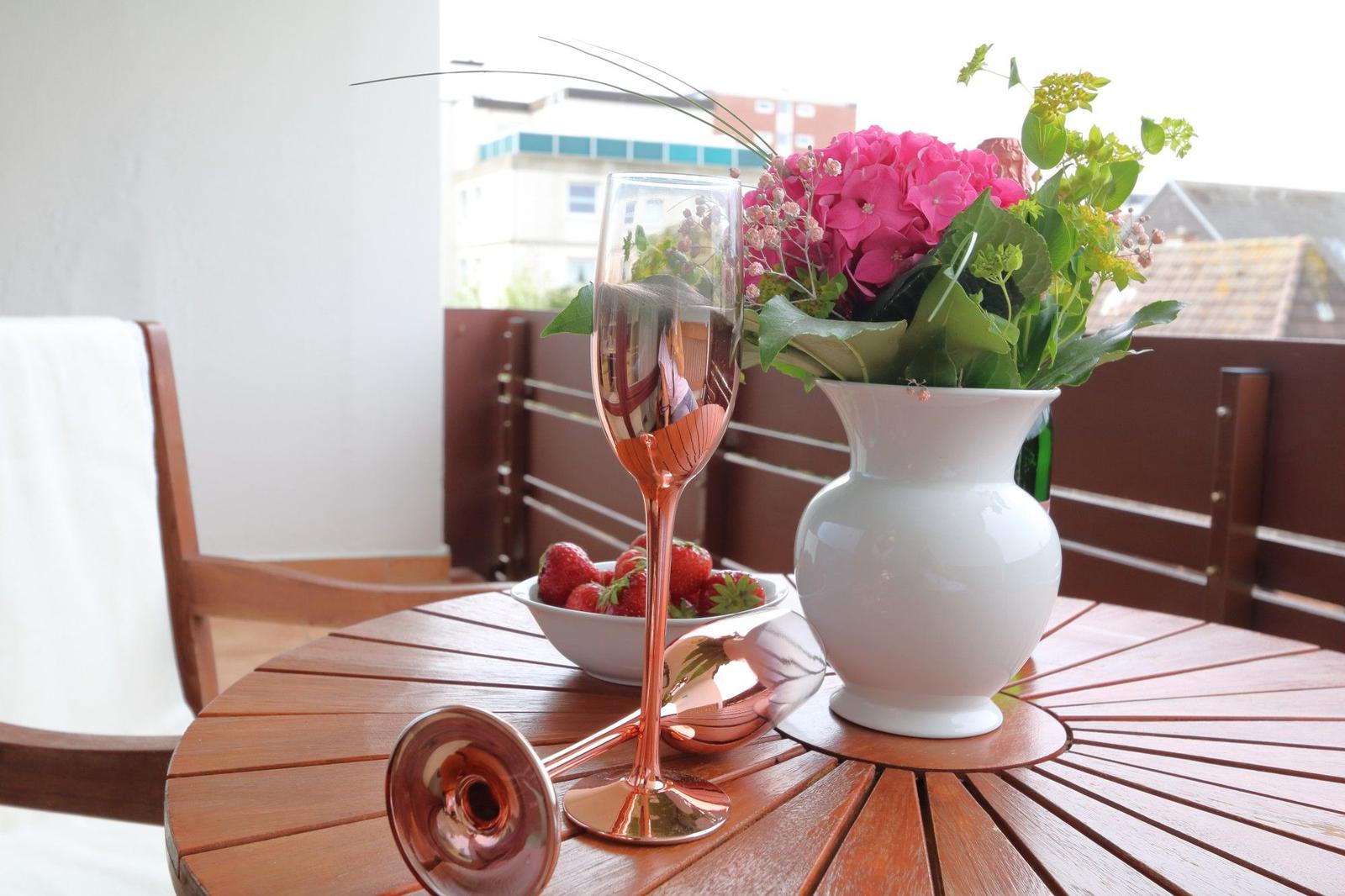 Auf der Terrasse steht ein runder Tisch mit Blumen, einem Glas und einer Schale Erdbeeren.
