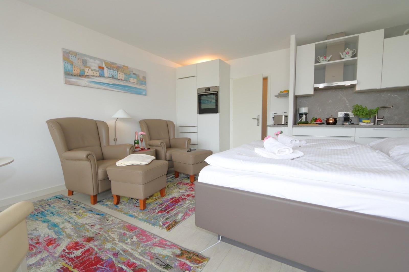 Schlafzimmer mit Bett, Küchenbereich und zwei Sesseln auf Teppich.