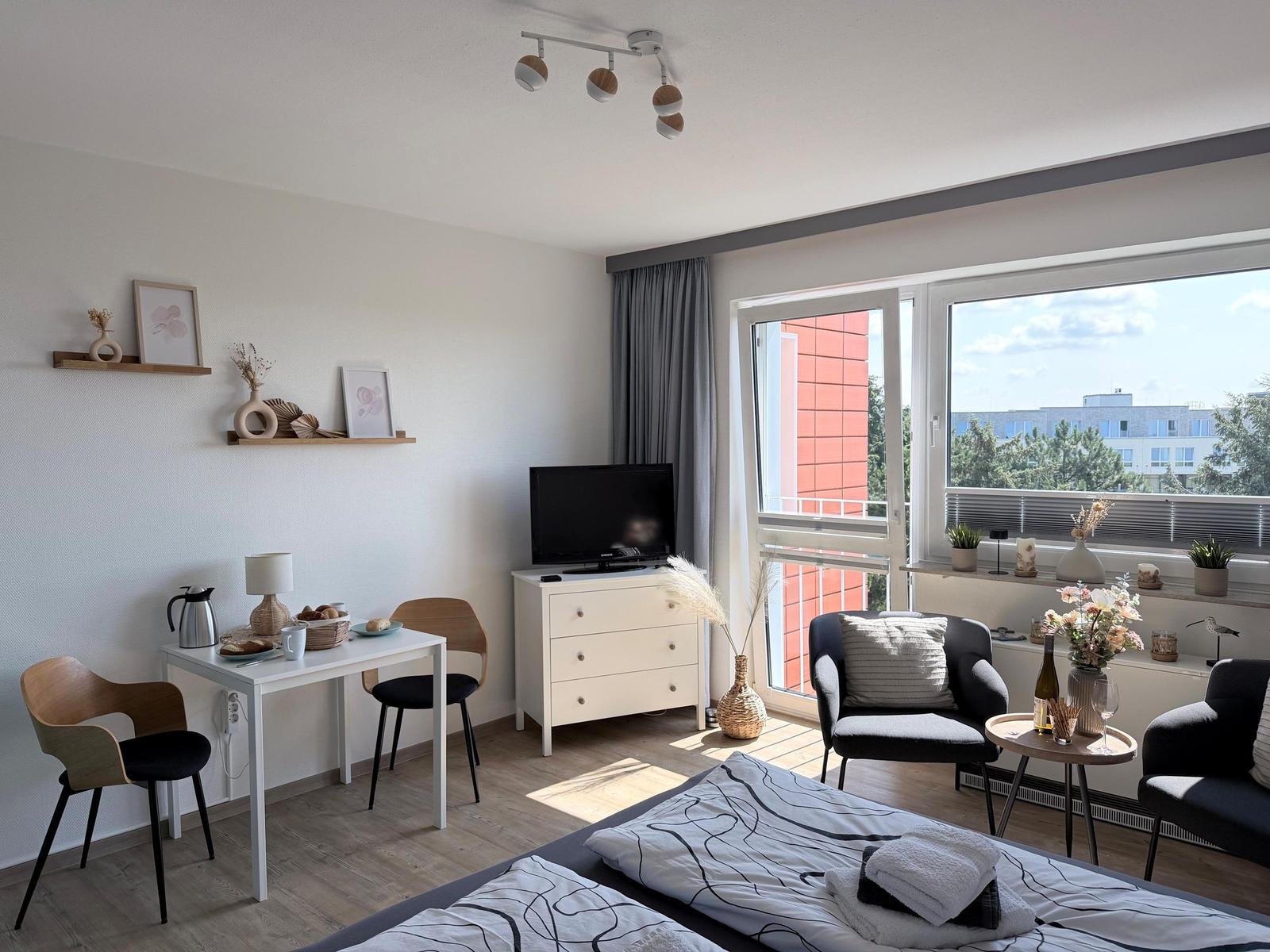 Zimmer mit Bett, Esstisch, TV und Balkon mit Blick auf Gebäude.