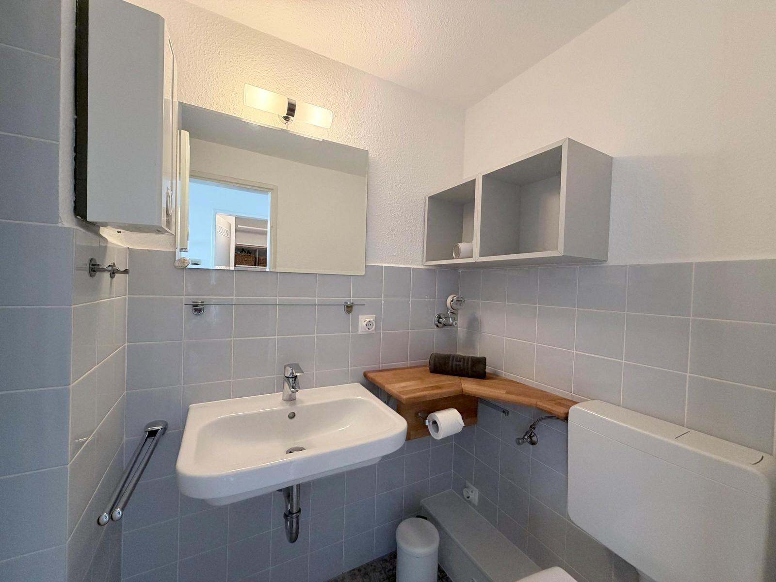 Badezimmer mit Waschbecken, Spiegel, Toilette und Holzablage.