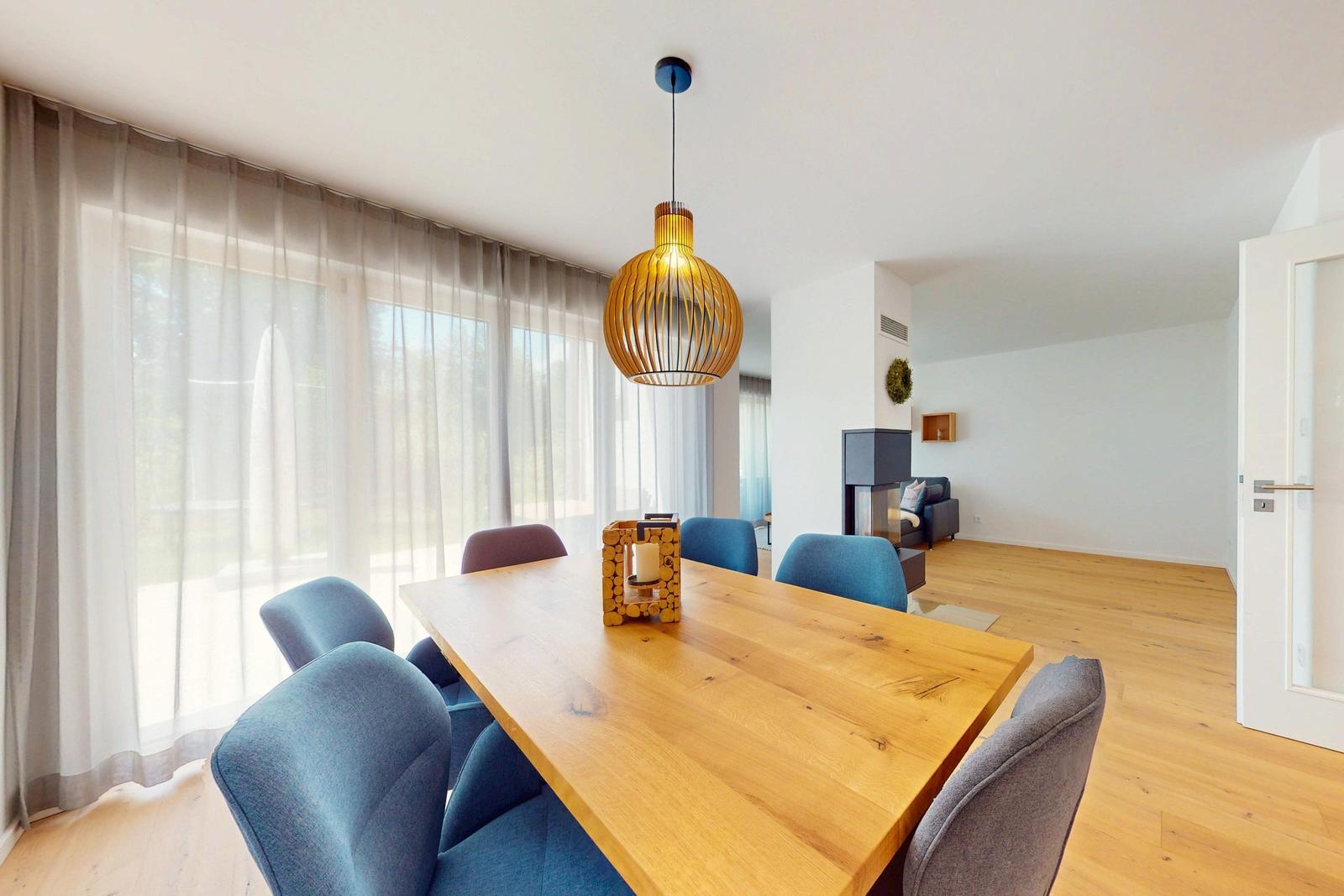 Esszimmer mit Holztisch, blauen Stühlen und Deckenleuchte. Durchgehende Fensterfront mit Gardinen.