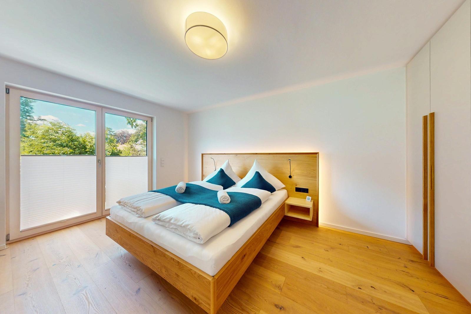 Schlafzimmer mit Holzbett, Fenster und blauer Bettwäsche.