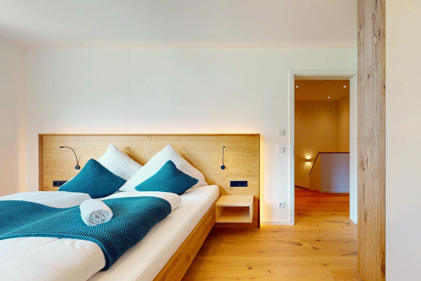 Schlafzimmer mit Bett, Holzrahmen und blauer Decke. Blick in angrenzenden Raum.