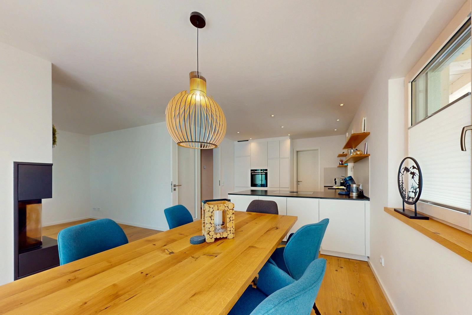 Essbereich mit Holztisch, blauen Stühlen und einer Deckenlampe. Im Hintergrund Küche und Kamin.